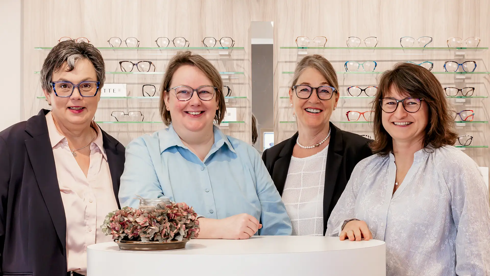 Das Team von Uhren Optik Arnold Stange in Bünde stehen lächelnd im Brillenfachgeschäft vor einer Wand mit Brillenfassungen